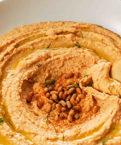 A hand dips a pita chip into a low bowl of orange hummus.