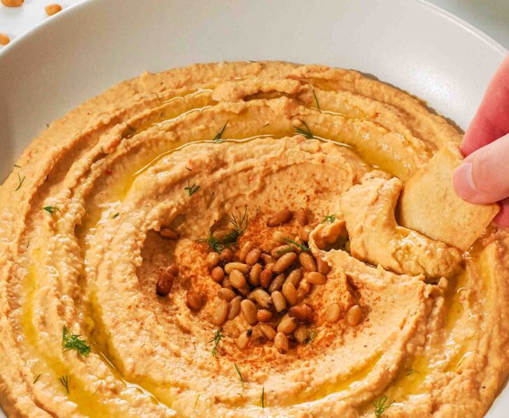 A hand dips a pita chip into a low bowl of orange hummus.