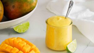 A small jar of mango curd with a lime wedge and slice of mango nearby.