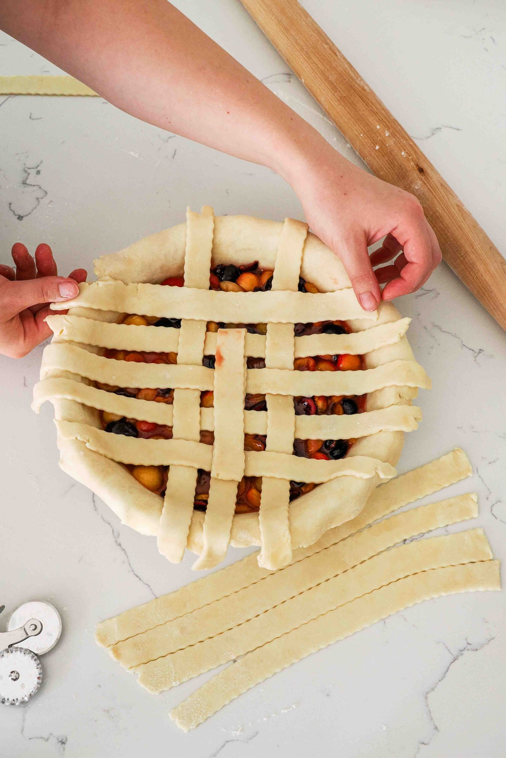 Two hands weave pie dough strips in a lattice over cherry pie filling.