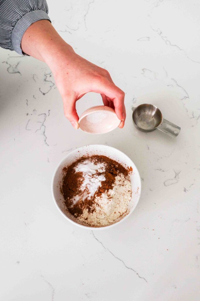A hand adds baking powder, baking soda, and salt to a bowl with flour and cocoa powder.
