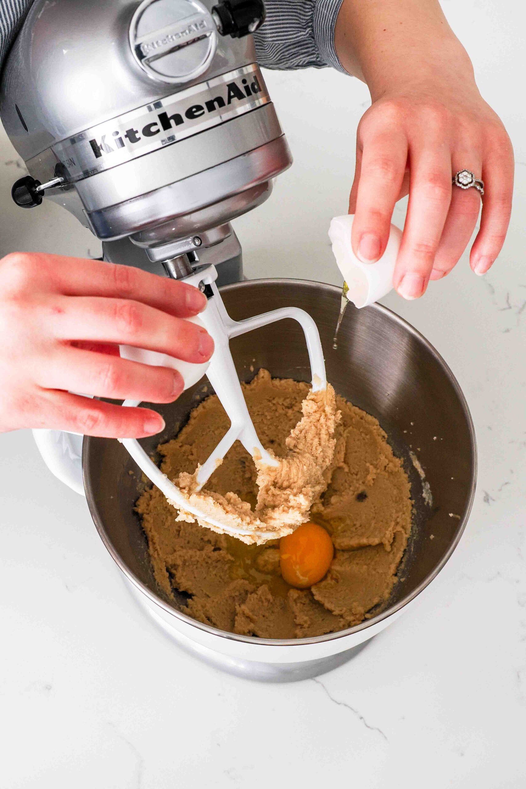 Two hands crack an egg into creamed butter and sugar in a mixer bowl.