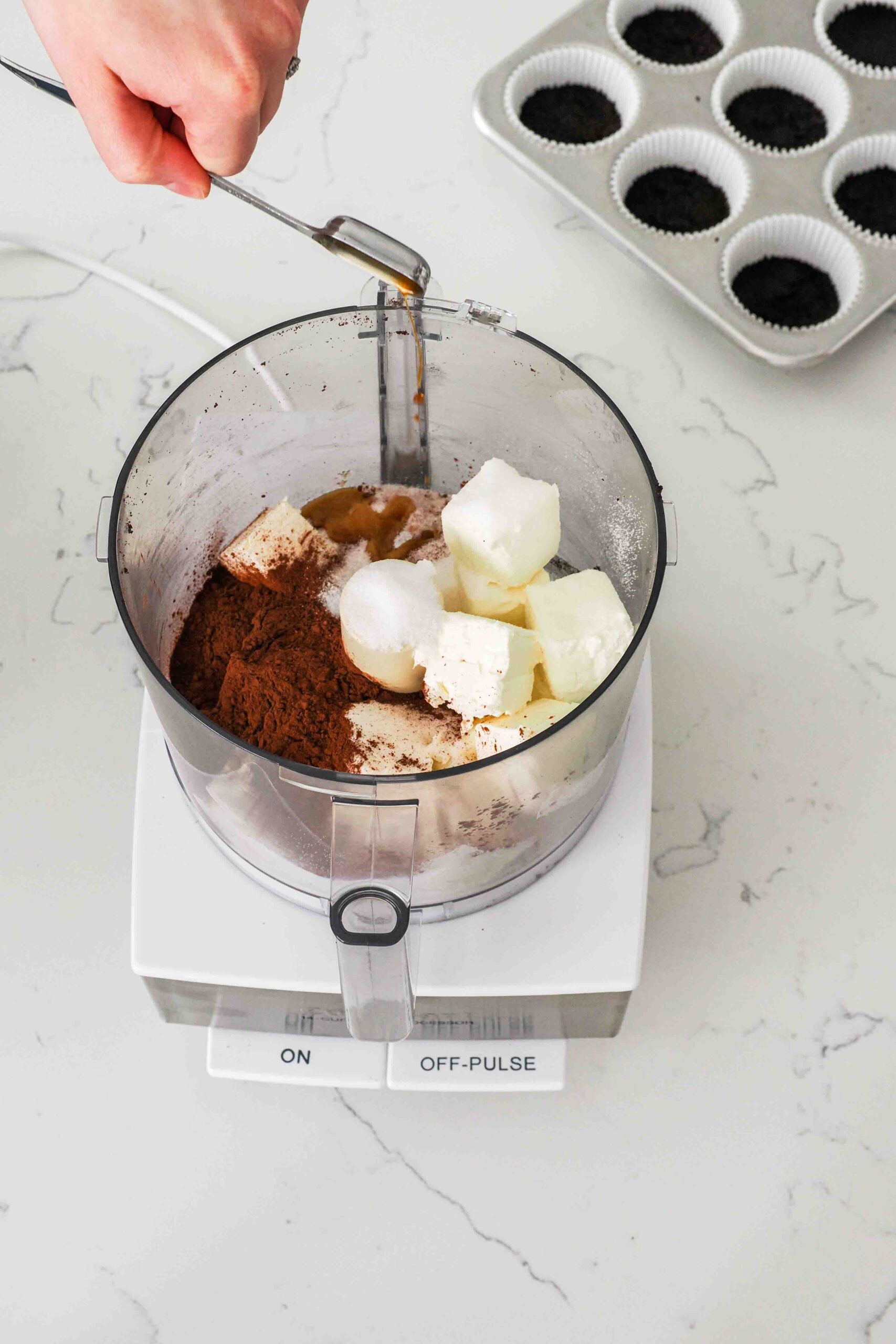 Vanilla is being added to a food processor bowl with other ingredients for a chocolate cheesecake.