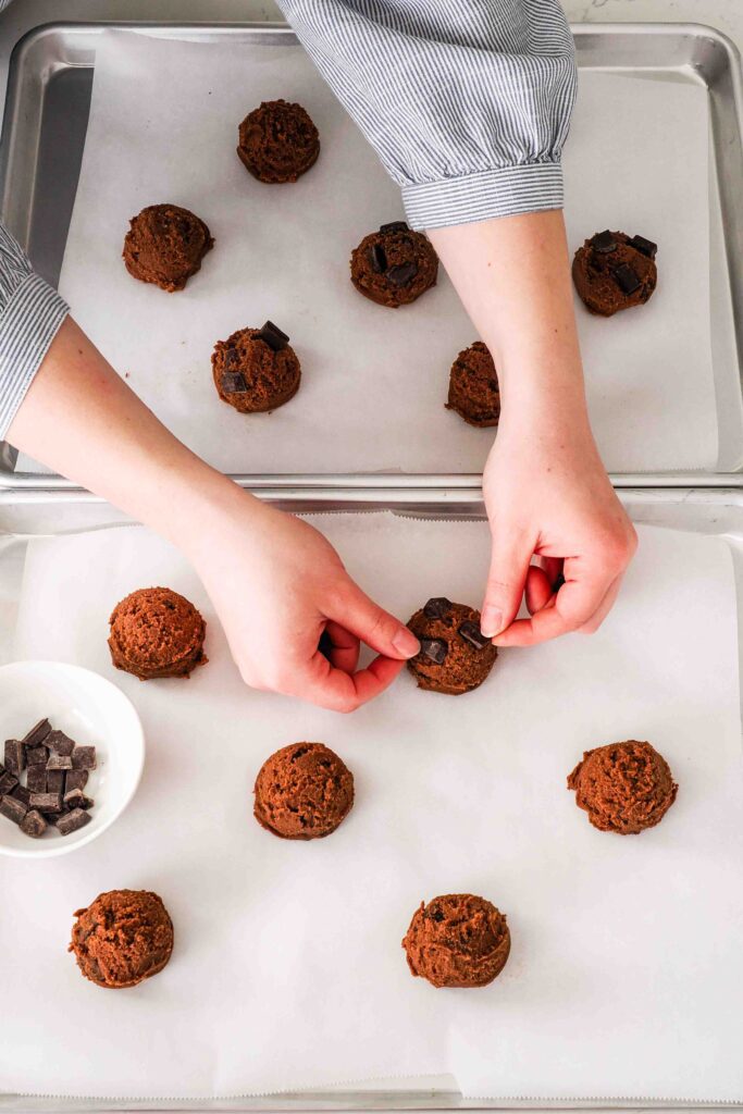 Two hands push more chocolate chips onto cookie dough balls on baking sheets.