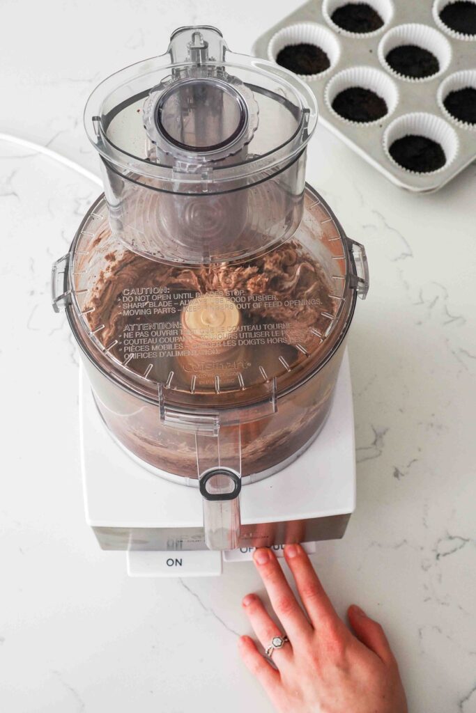 A hand presses "pulse" on a food processor to mix chocolate cheesecake ingredients.