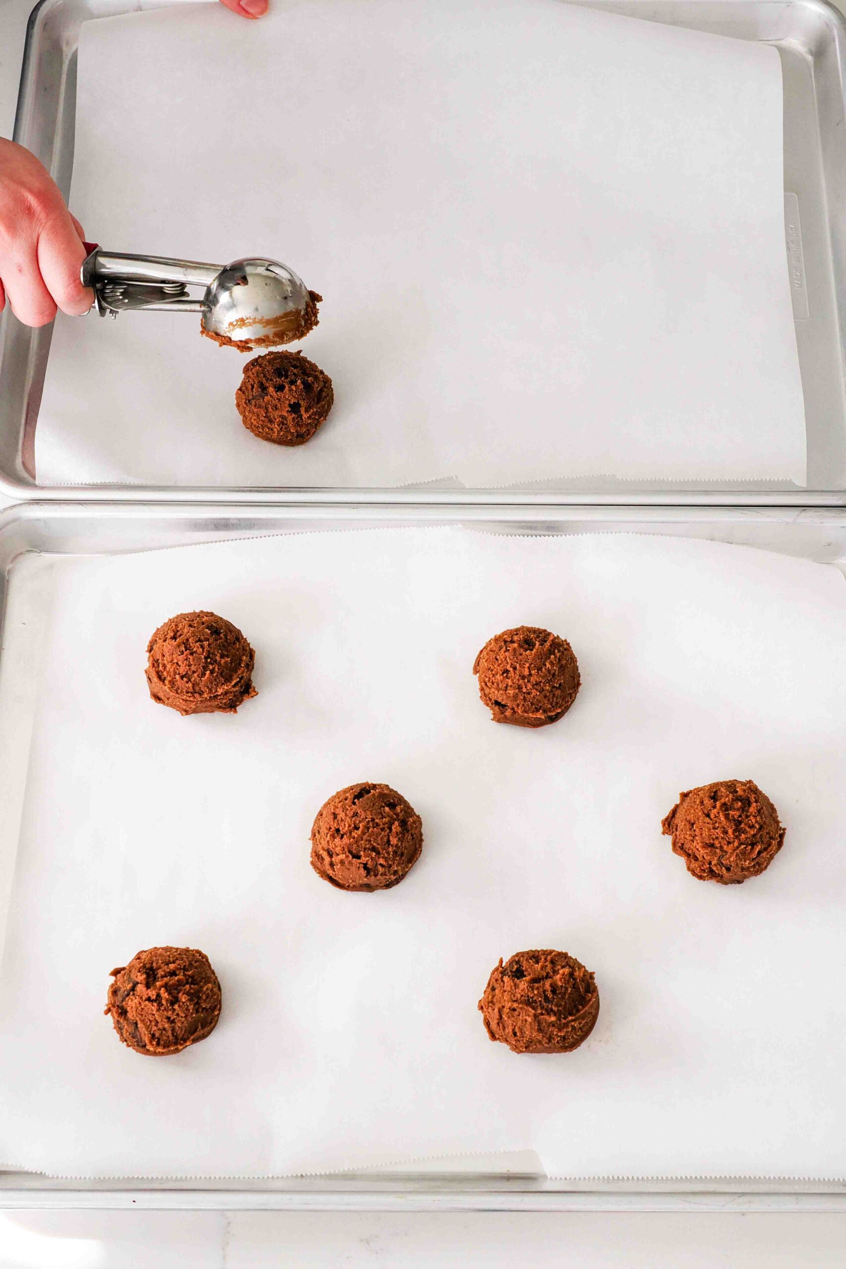 A hand scoops 2" balls of chocolate cookie dough onto parchment-lined baking sheets.