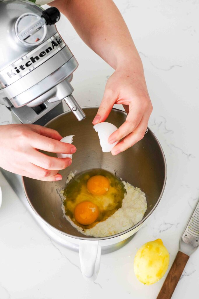 Two eggs are cracked into a stand mixer bowl with lemon sugar.