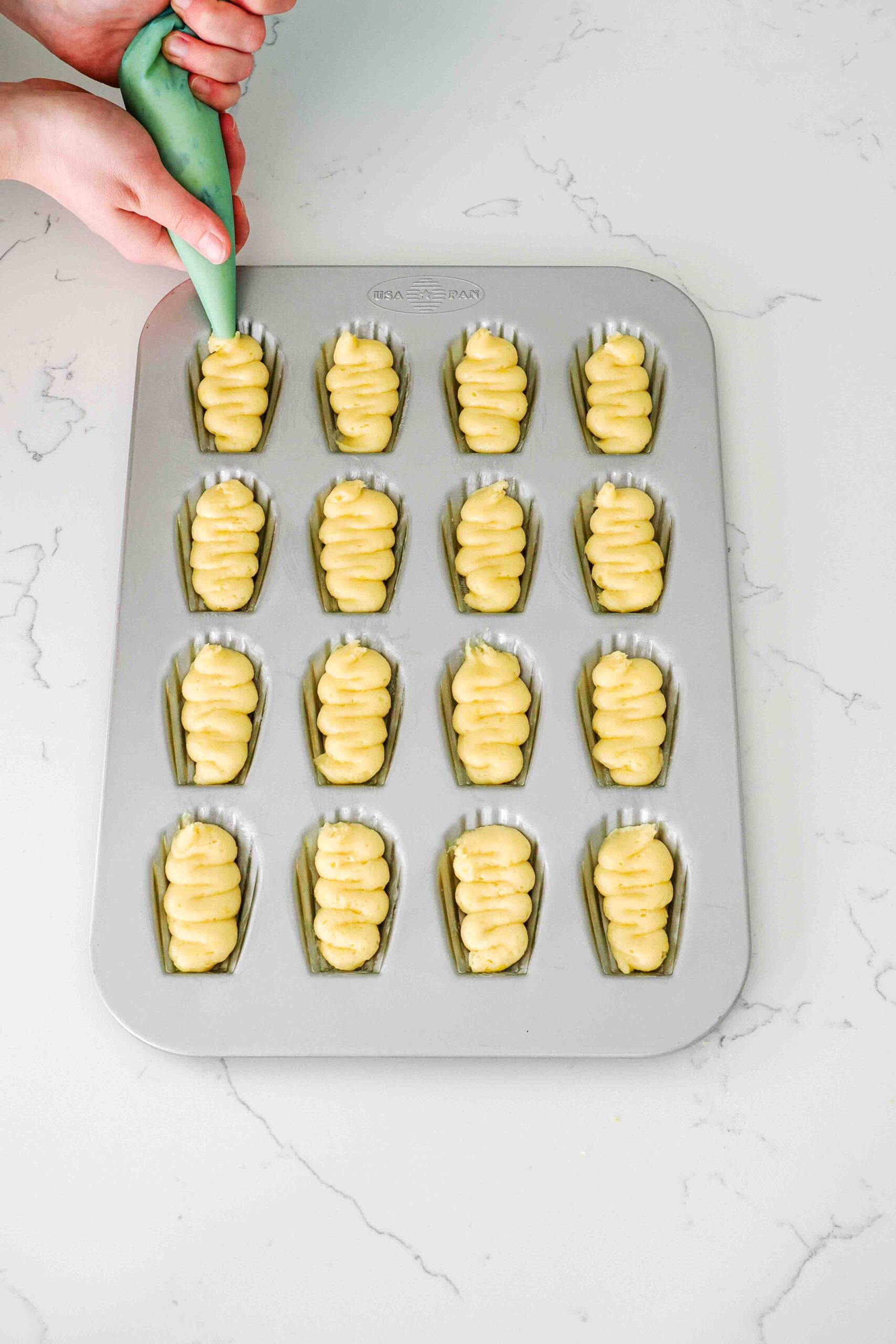 A hand pipes lemon madeleine batter into a madeleine pan.