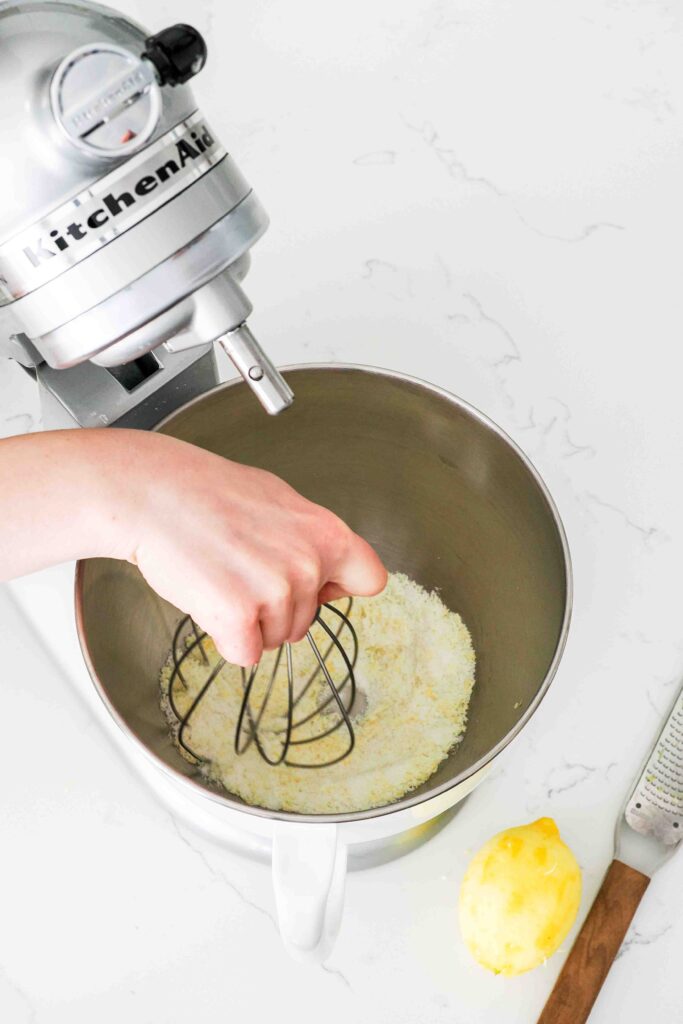 A hand uses the whisk on a KitchenAid mixer to stir together lemon zest and sugar.