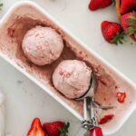 Two scoops of strawberry ice cream in a container with strawberries nearby.