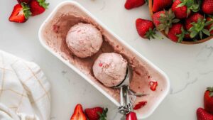 Two scoops of strawberry ice cream in a container with strawberries nearby.