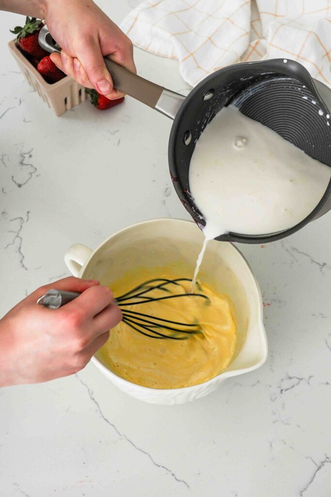 Warm cream is poured from a pot into a mixing bowl with ribboned egg yolks.