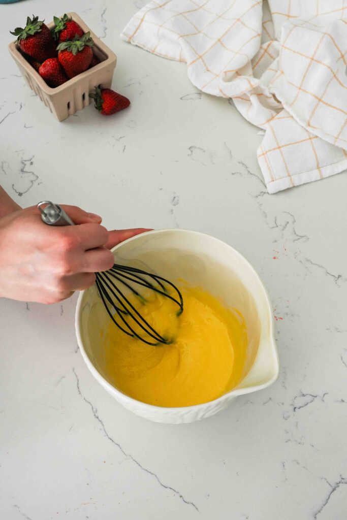 A hand whisks together sugar and egg yolks in a mixing bowl.