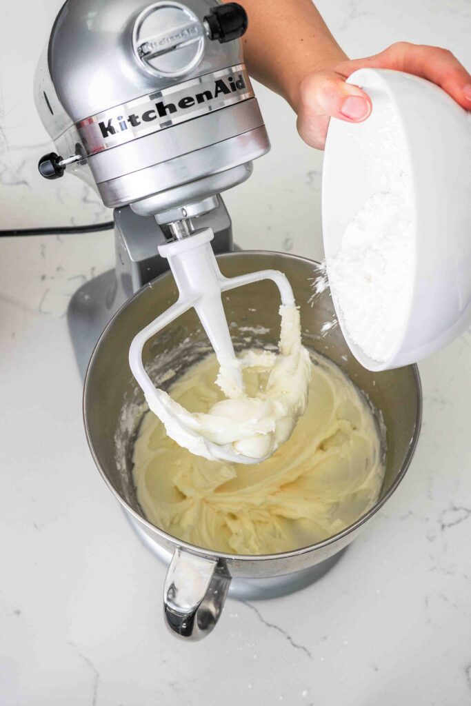 Powdered sugar is poured into a mixing bowl with creamed butter.