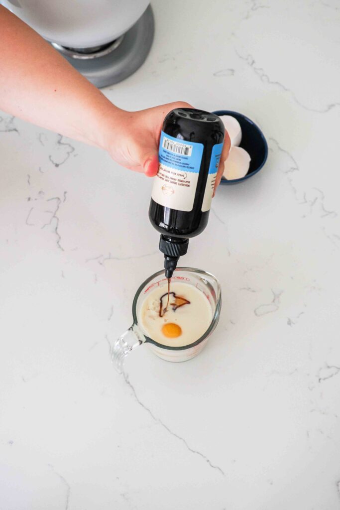 A hand squeezes vanilla bean paste into a measuring cup with milk and eggs.