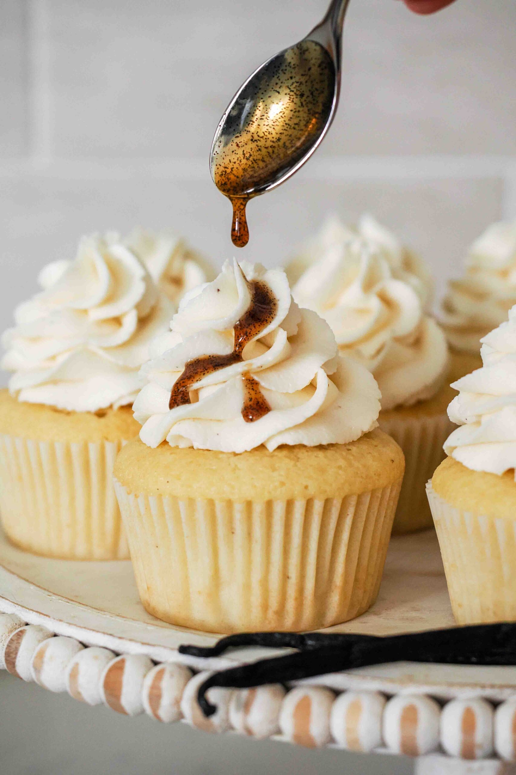 A spoon drizzles vanilla bean paste over a vanilla cupcake.