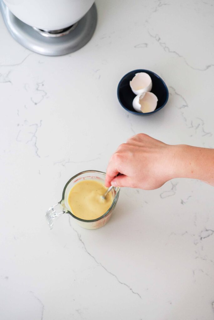 A hand whisks eggs, milk, and vanilla bean paste together in a measuring cup.