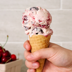 A hand holds up a scoop of black cherry ice cream in a cone.