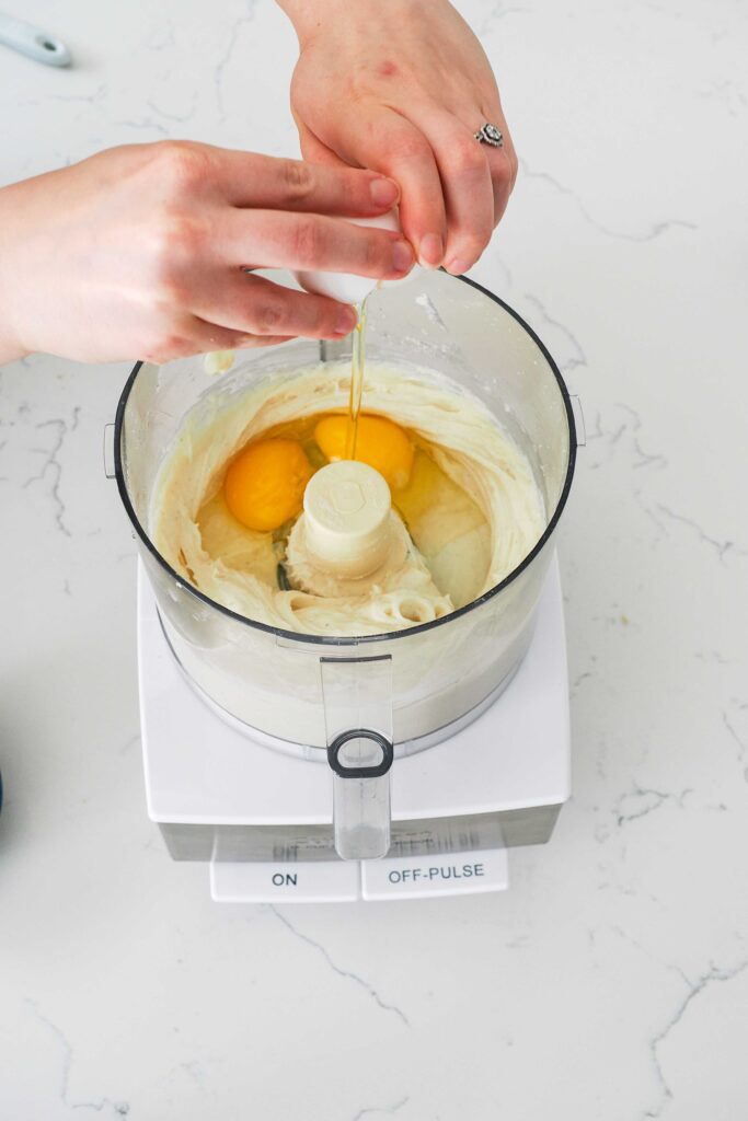 Eggs are added to a food processor bowl with blended cream cheese and sugar.