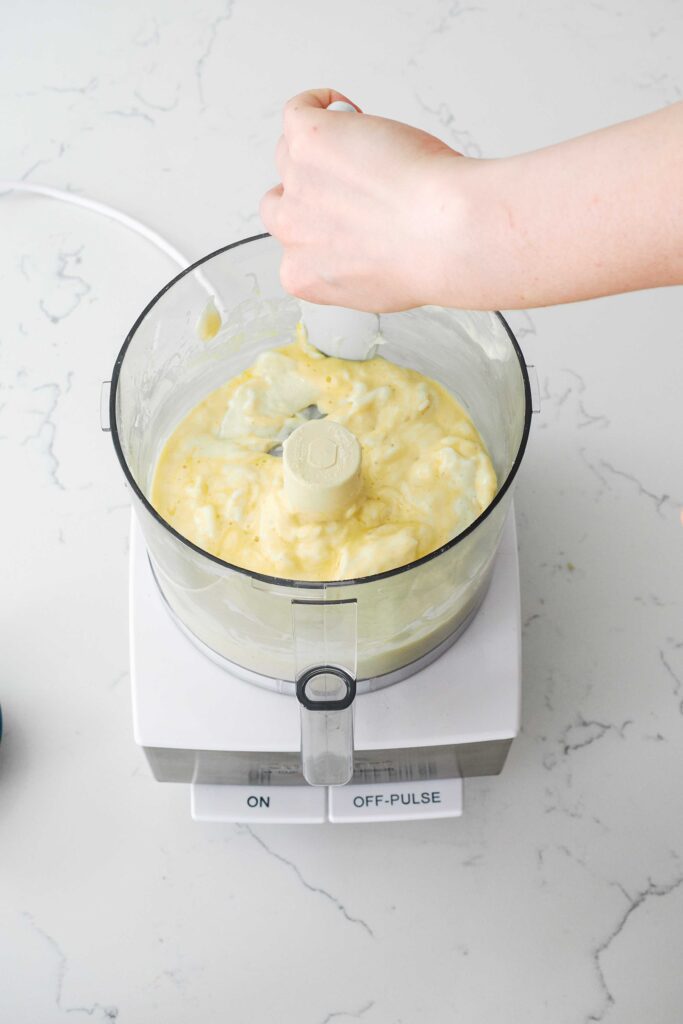 A hand guides a spatula around the bowl of a food processor with lumpy cheesecake filling.
