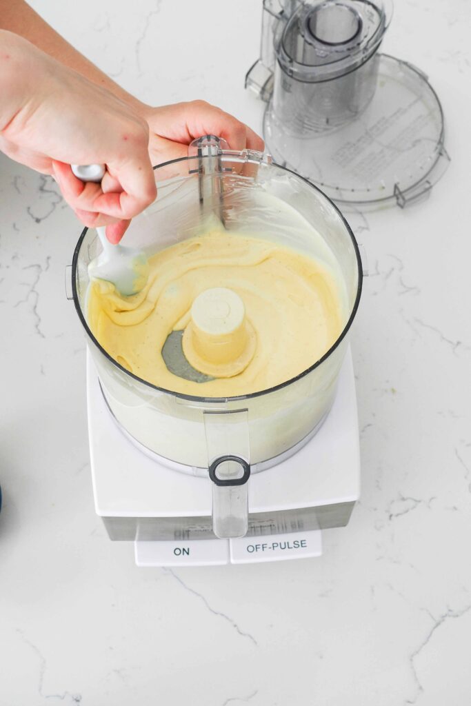 A hand guides a spatula around the edge of the bowl of a food processor with a smooth cheesecake filling.