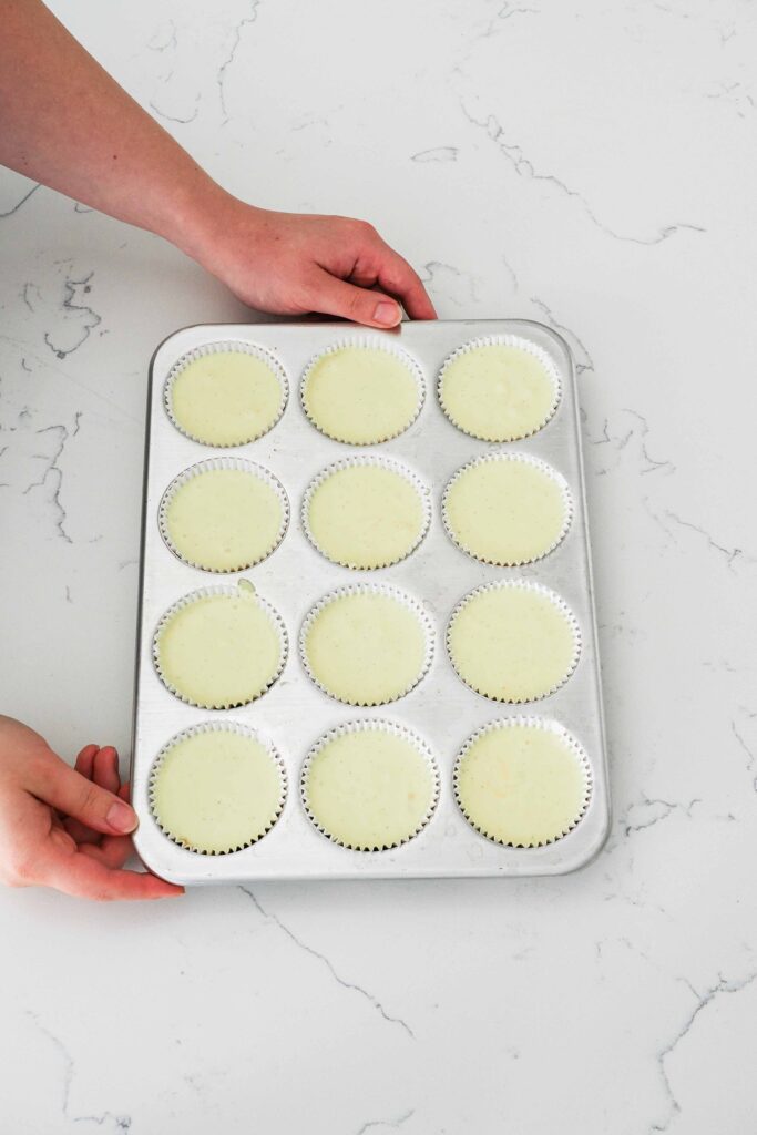 Two hands hold a muffin pan of unbaked vanilla cheesecakes.