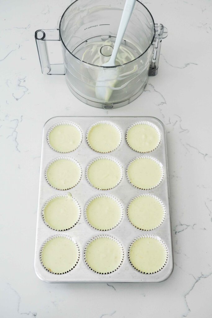 An empty food processor bowl next to a muffin pan with unbaked mini cheesecakes.