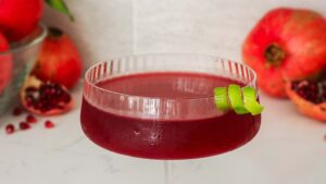 A coupe glass filled with a pomegranate cosmopolitan and garnished with a lime twist.