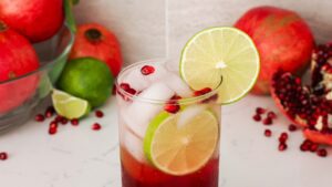 A tall glass with an iced pomegranate mojito is topped with a lime wheel and pomegranate arils.