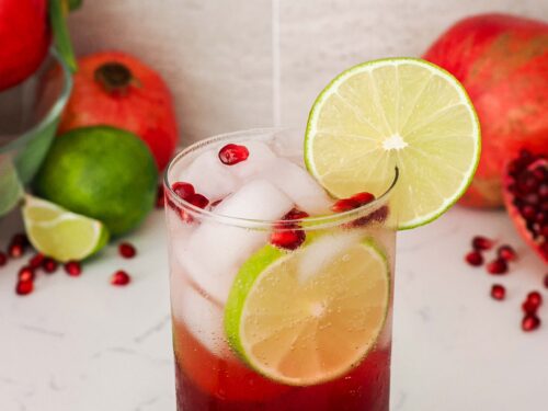 A tall glass with an iced pomegranate mojito is topped with a lime wheel and pomegranate arils.