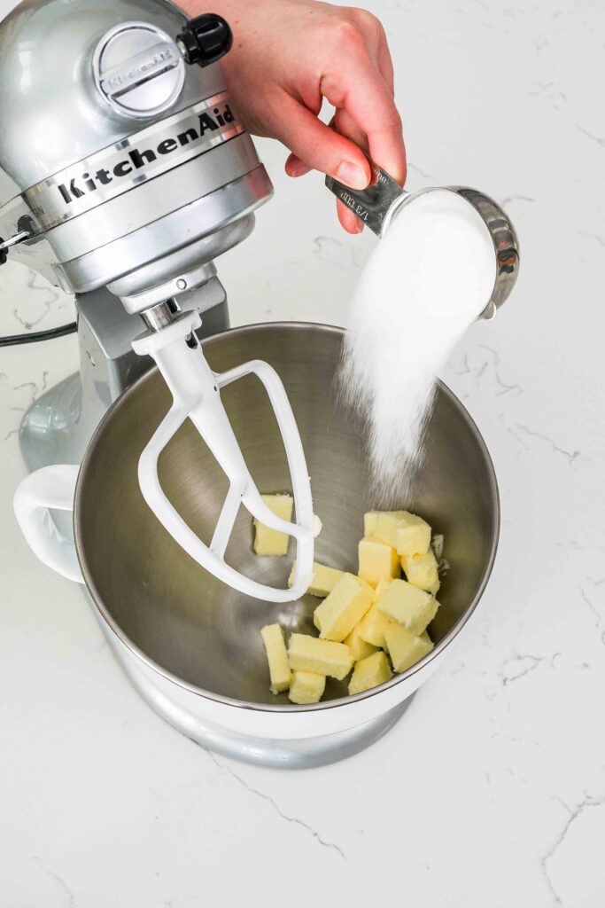 A hand pours granulated sugar into a mixing bowl with butter.