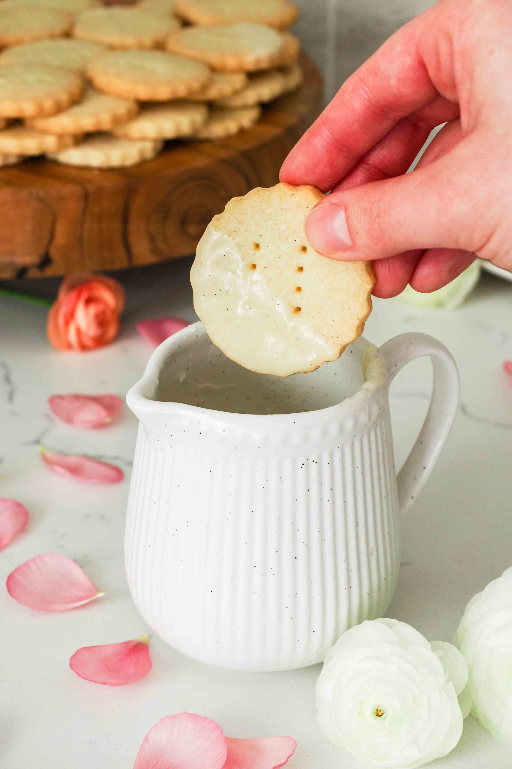A hand dips a vanilla shortbread cookie into a small jug of vanilla glaze.