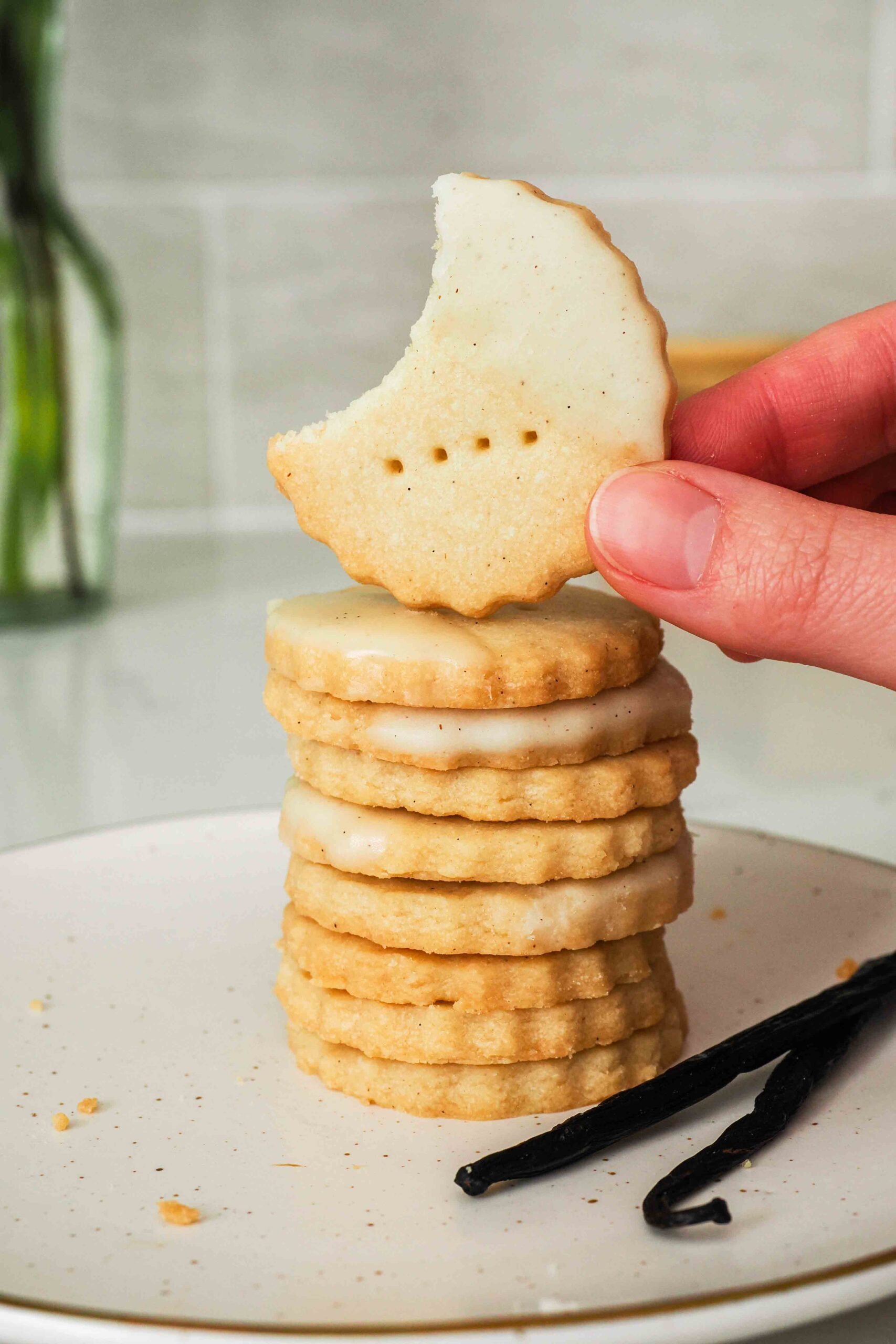 A hand holds up a vanilla shortbread cookie missing a bite in front of a stack of cookies.