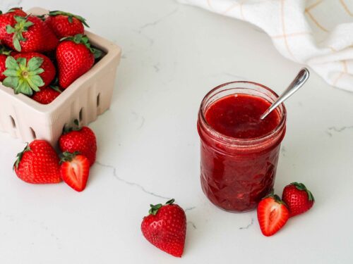 A jar of strawberry jam near a basket of strawberries.