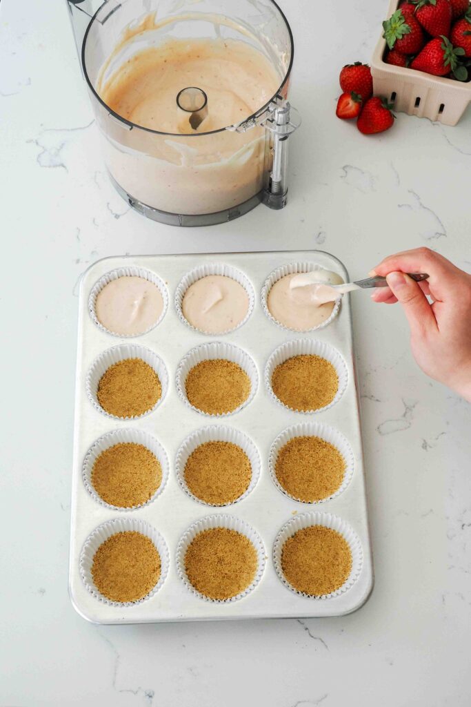 Strawberry cheesecake batter is added on top of graham cracker crusts in a muffin pan.