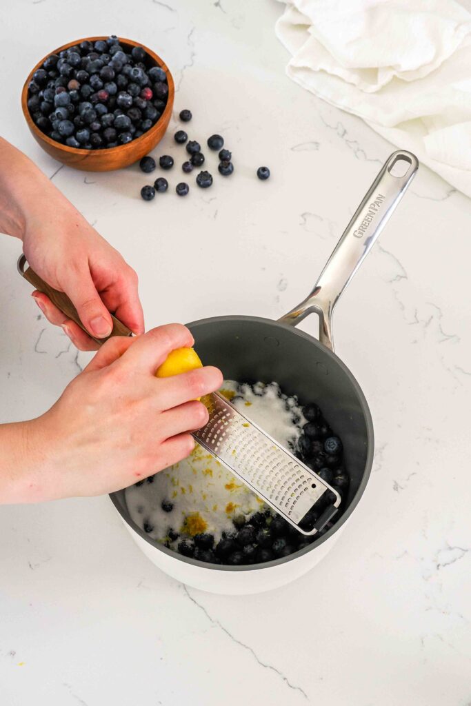Lemon is zested over a pot of blueberries and sugar.